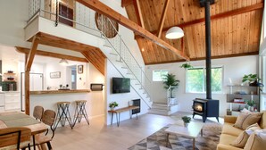 A spacious living area with a high ceiling, wooden beams, a fireplace, a staircase, and a kitchen area with bar stools. - Modern A-Frame next to a creek, minutes to Bass Lake and close to Yosemite  (Oakhurst)