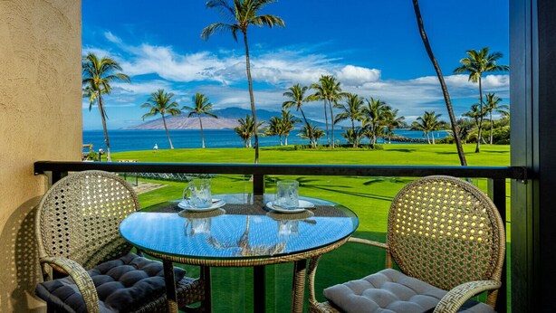 A balcony with a glass table and wicker chairs, overlooking a golf course and ocean. - Enjoy Your Oceanside Dream at Kihei Surfside (Kihei)