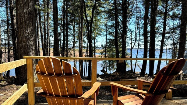 Una terraza de madera con dos sillas Adirondack mirando a un lago rodeado de árboles.