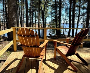 Eine Holzterrasse mit zwei Adirondack-Stühlen, die auf einen See mit Bäumen blickt.