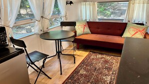 Un espacio habitable compacto con un sofá, una mesa de comedor y una silla. Hay dos ventanas con cortinas, un reloj en la pared y una alfombra pequeña.
