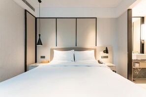 A modern hotel room with a large bed, bedside tables, a nightstand, a wall-mounted lamp, and a bathroom visible through an open door. - Ji Hotel (Xiamen)