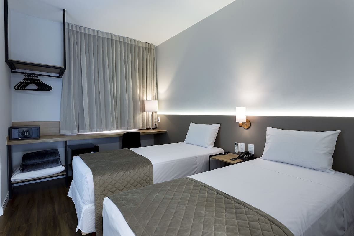 A hotel room with two beds, a desk, a chair, a lamp, a radio, and a coat rack.