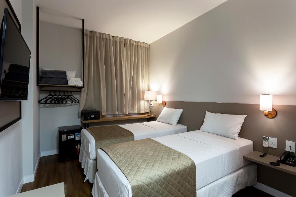 A hotel room with two beds, a bedside table with a lamp, a wardrobe, and a mirror.