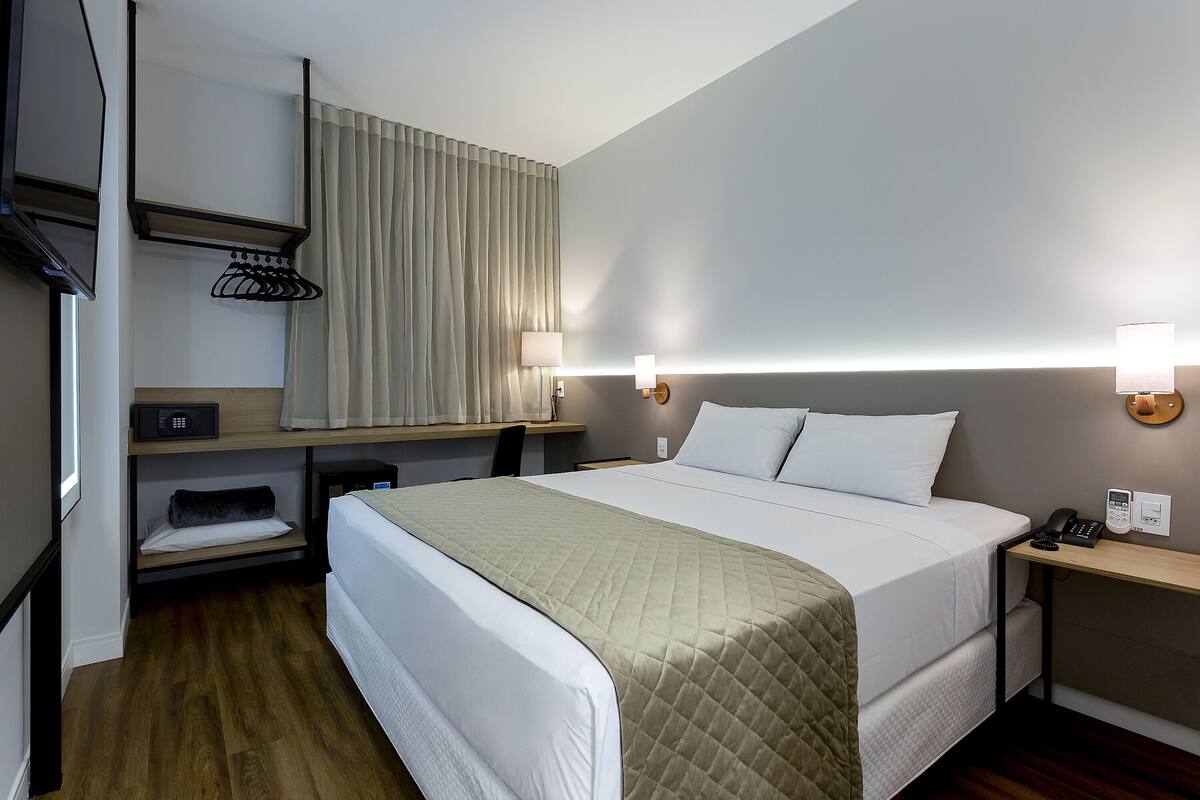 A hotel room with a bed, bedside tables, a desk, a chair, a TV, a coat rack, and a window with curtains.