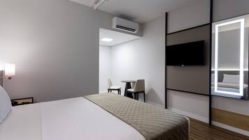 Et hotelværelse med en seng, et fjernsyn ophængt på væggen, et lille bord og stole samt en aircondition.