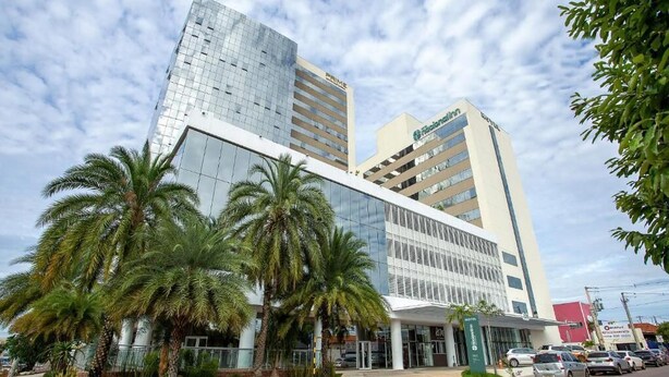 A modern hotel with a glass facade, palm trees in the front, and a parking lot with cars. - Nacional Inn Rondonopolis (Rondonópolis)