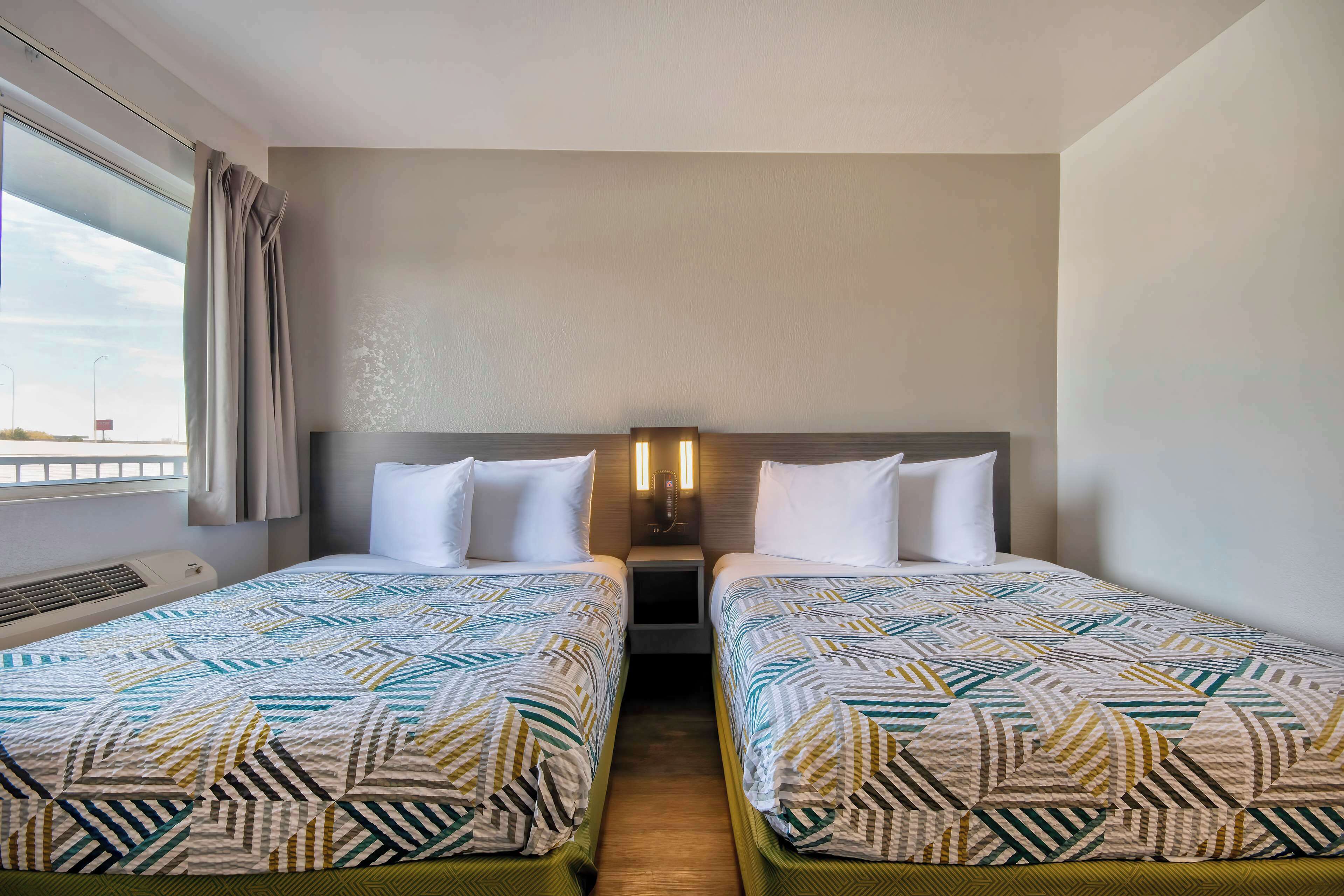 Quarto de hotel com duas camas, janela com cortinas, luminária fixada na parede e uma pequena mesa de cabeceira.