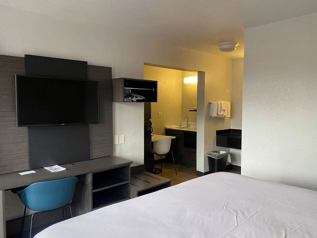 A hotel room with a bed, a desk with a chair, a TV mounted on the wall, a small bathroom with a sink and mirror, and a trash bin.