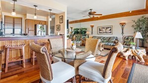 A spacious living area with a glass-top coffee table surrounded by wicker chairs, a wooden dining table with bar stools, and a cozy seating area with patterned cushions. - Private Oceanfront Penthouse Lae Nani 231, New split A/C, Parking, AMAZING VIEW! (Kapaʻa)