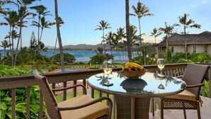 A round glass table with wicker chairs on a balcony overlooking a beach with palm trees and a view of the ocean. - Private Oceanfront Penthouse Lae Nani 231, New split A/C, Parking, AMAZING VIEW! (Kapaʻa)