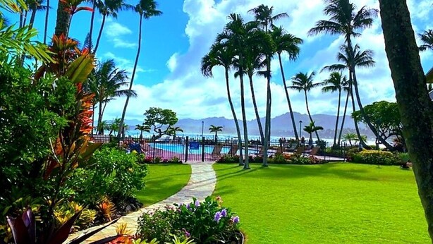 A tropical garden with palm trees, a pathway, and a view of the ocean. - Private Oceanfront Penthouse Lae Nani 231, New split A/C, Parking, AMAZING VIEW! (Kapaʻa)