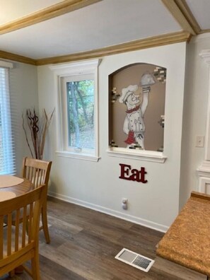 A dining area with a wooden table and chairs, a window with a view of trees, and a wall-mounted artwork of a chef.