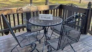 Een houten terras met een metalen tafel en stoelen, een bordje met de tekst 'wees onze gast', en een uitzicht op bomen en een weg in de verte.