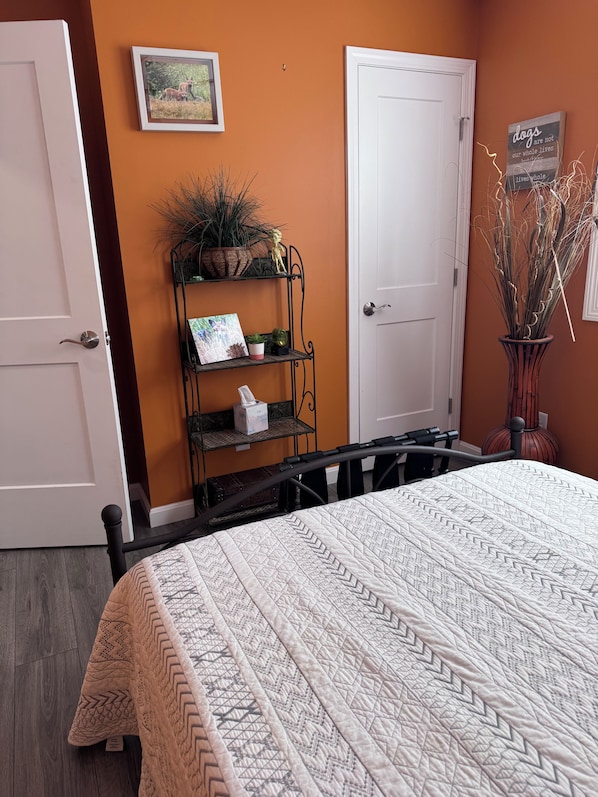 A bedroom with a bed, a shelf with plants, a door, and a wall with a framed picture.