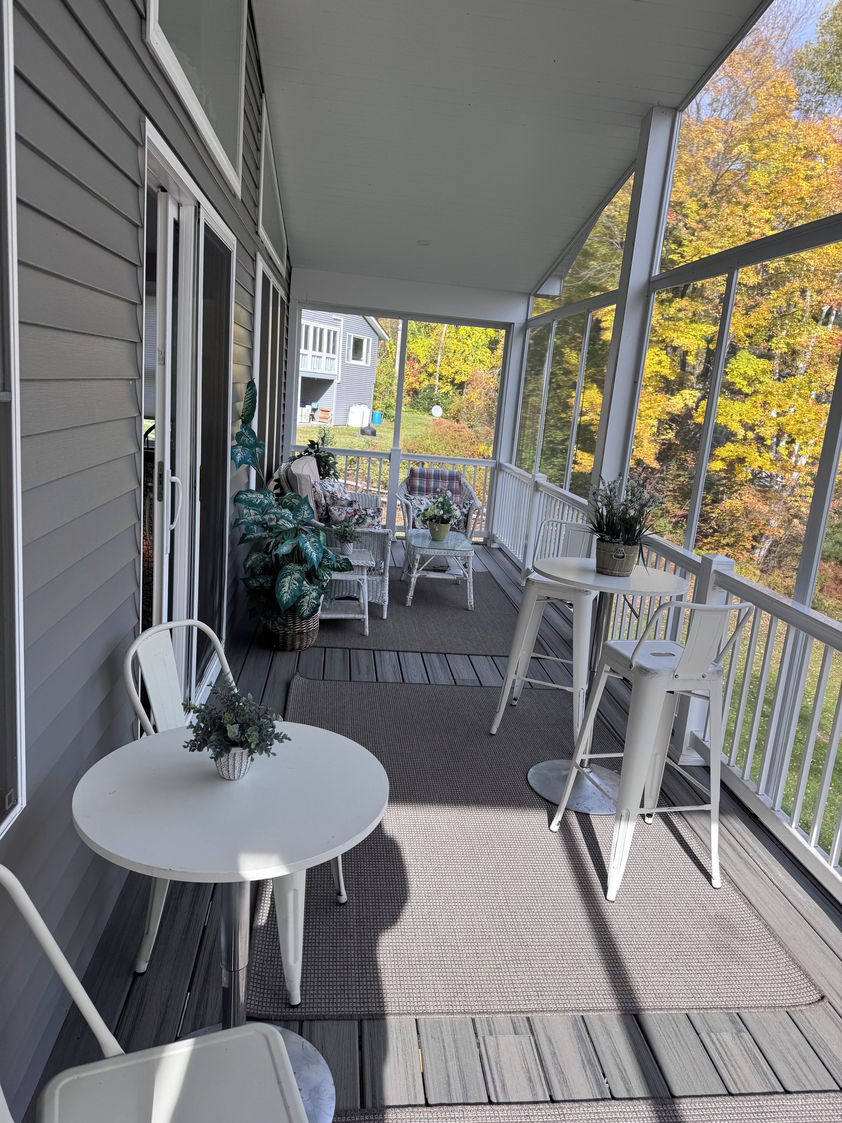 Een overdekt terras met witte stoelen en tafels, een uitzicht op een huis, en herfstkleurige begroeiing.