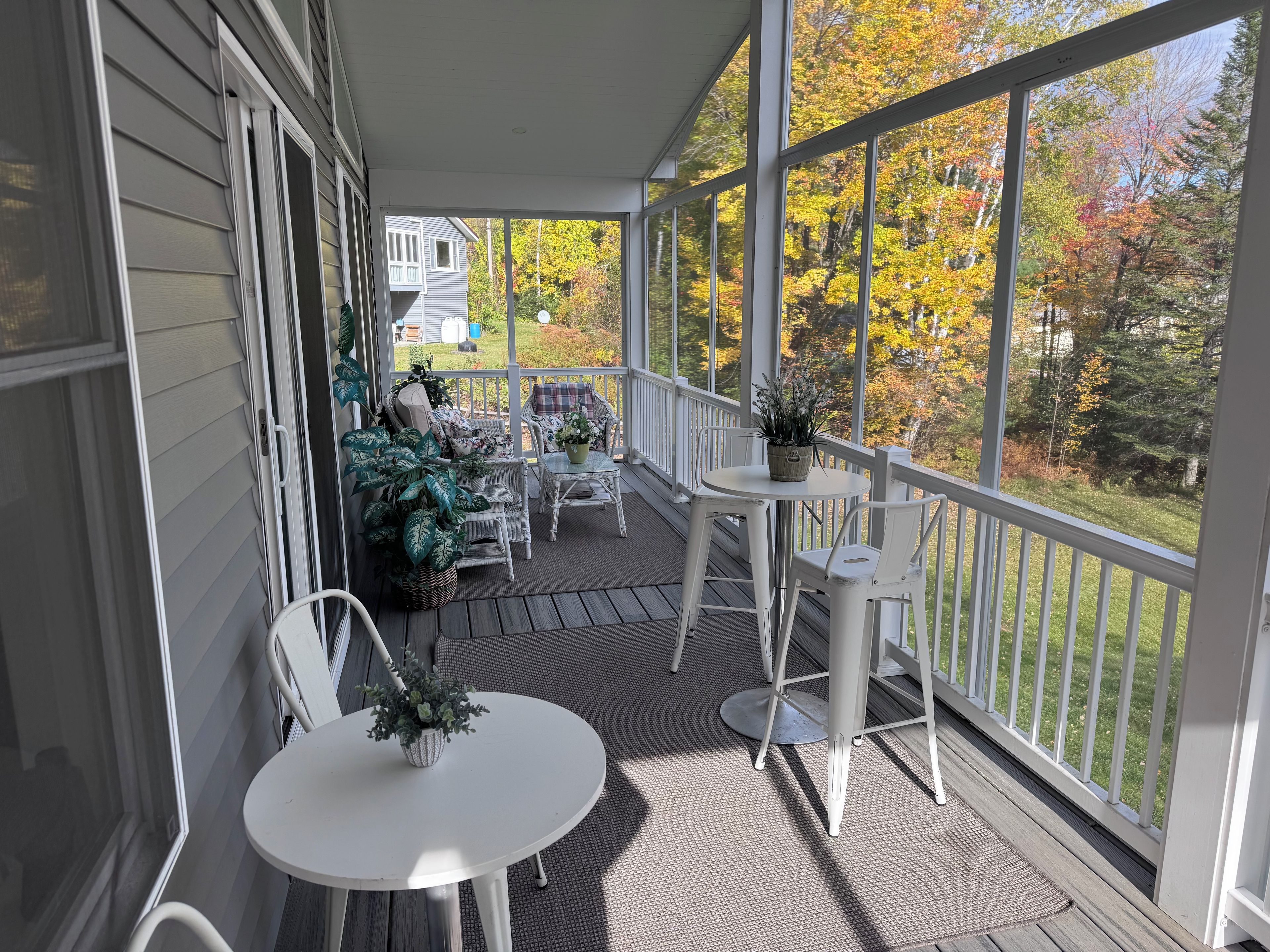 A covered porch with outdoor furniture, potted plants, and a view of a house and autumn trees.