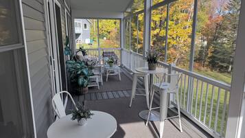 A covered porch with outdoor furniture, potted plants, and a view of a house and autumn trees.