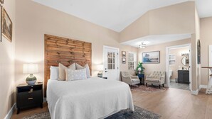 A bedroom with a large bed, a wooden headboard, a nightstand with a lamp, a chair, and a view into another room. - COZY COTTAGE - Private Hot Tub and Fire Pit (Branson)