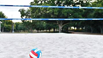 A volleyball on a sandy surface with a net and trees in the background.