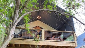 Een houten terras met een metalen leuning, een overdekt gebied met een zwart dak, en een boom op de voorgrond.