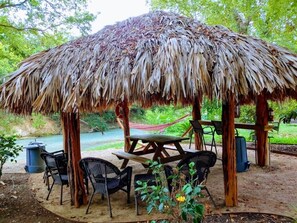 A thatched-roof pavilion with a picnic table and chairs, surrounded by greenery and a river. - Charming Cabin on the San Marcos River, comfortable, clean, fun, lots to do! L4 (Kingsbury)