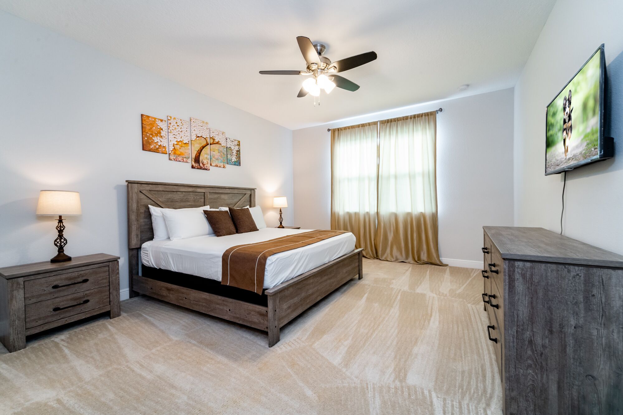 A bedroom with a wooden bed, nightstands, a ceiling fan, a large window with curtains, and a TV on the wall.