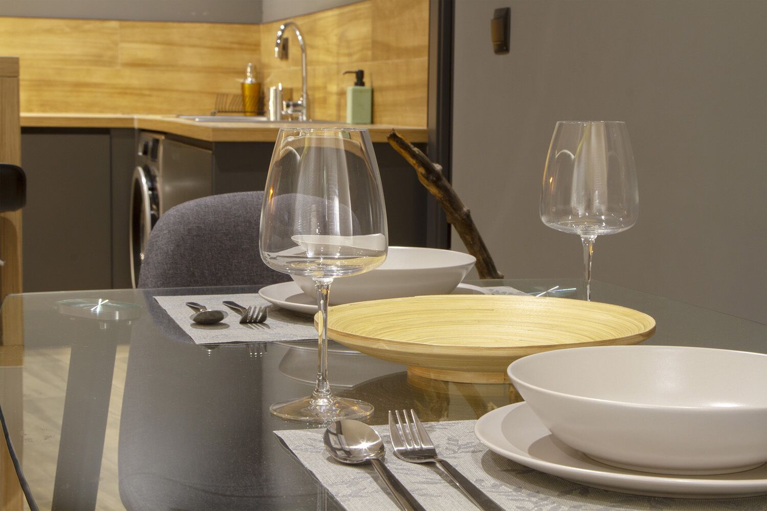 Un moderno juego de mesa con platos blancos, tazones y copas de vino transparentes, un salero de madera y una cuchara sobre un mantel individual.