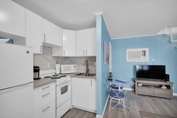 A compact kitchen with white cabinets, a microwave, a stove, and a sink. There's a small dining area with a chair and a TV on a wooden stand.