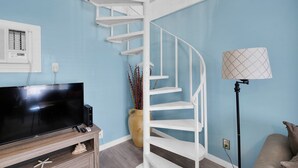 Ein Zimmer mit Wendeltreppe, einem Flachbildfernseher auf einem Holzbalken, einer Stehlampe und einer Topfpflanze.