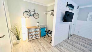 Ein Zimmer mit Holzboden, einem an der Wand montierten Fernseher, einem Fahrrad, einem kleinen Tisch mit Schubladen und einer Topfpflanze.