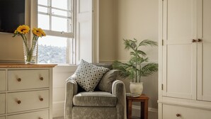A room with a grey armchair, a wooden side table with a potted plant, a dresser with drawers, and a window with a view.