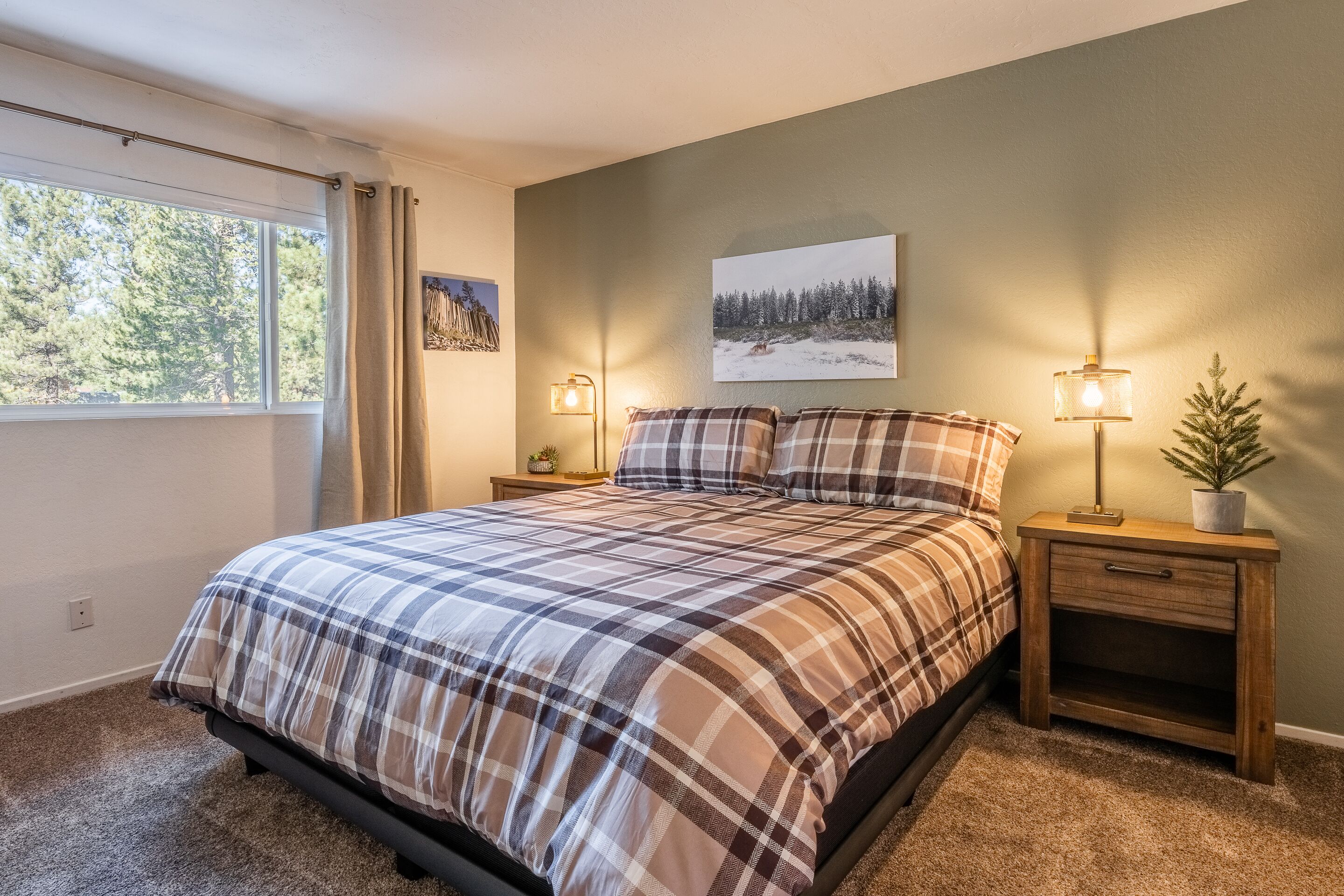 A bedroom with a large bed, two bedside tables with lamps, a window with curtains, and a wall-mounted painting.