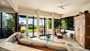 A spacious living area with a large sofa, a glass coffee table, and a view of a lush garden through large windows. - 🏖️Updated Ground Floor Mauna Lani Condo on the Golf Course (Kamuela)