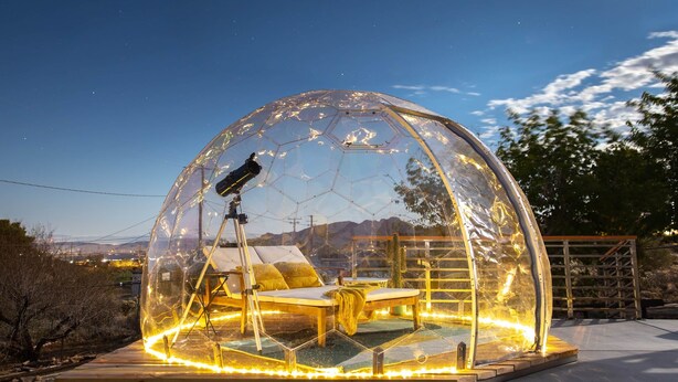 Een transparante koepelconstructie met een telescoop, een loungestoel en een tafel, omgeven door een houten terras en verlicht met lichtslingers.