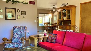 Una sala de estar con una mesa de centro de vidrio, un sillón estampado, un sofá rojo y un aparador de madera con vajilla.