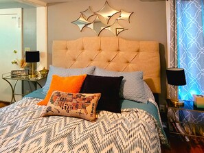A bedroom with a tufted headboard, a patterned bedspread, and multiple pillows. There is a side table with a lamp and a decorative mirror above the bed. - Jennywood Cottage - Prescott Cabin Rentals (Prescott)