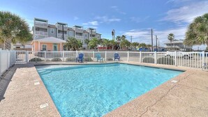 A swimming pool with a white fence, blue chairs, and a building with multiple floors in the background. - Blue Wave~Community Pool~Family Friendly~Sleeps 10 (Port Aransas)