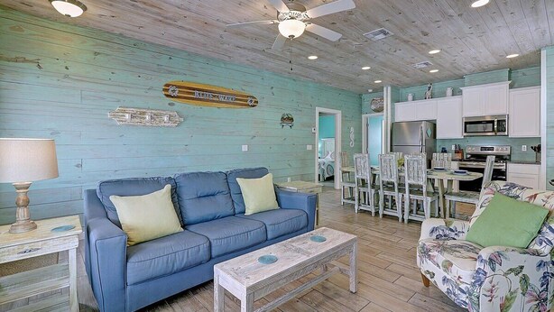 A cozy living room with a blue sofa, a floral armchair, and a wooden coffee table. A kitchen area with white cabinets and appliances is visible in the background. - Blue Wave~Community Pool~Family Friendly~Sleeps 10 (Port Aransas)