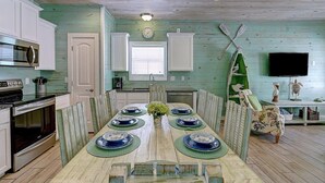 A dining area with a long table set for six, surrounded by chairs. There is a microwave and oven in the kitchen area to the left. - Blue Wave~Community Pool~Family Friendly~Sleeps 10 (Port Aransas)