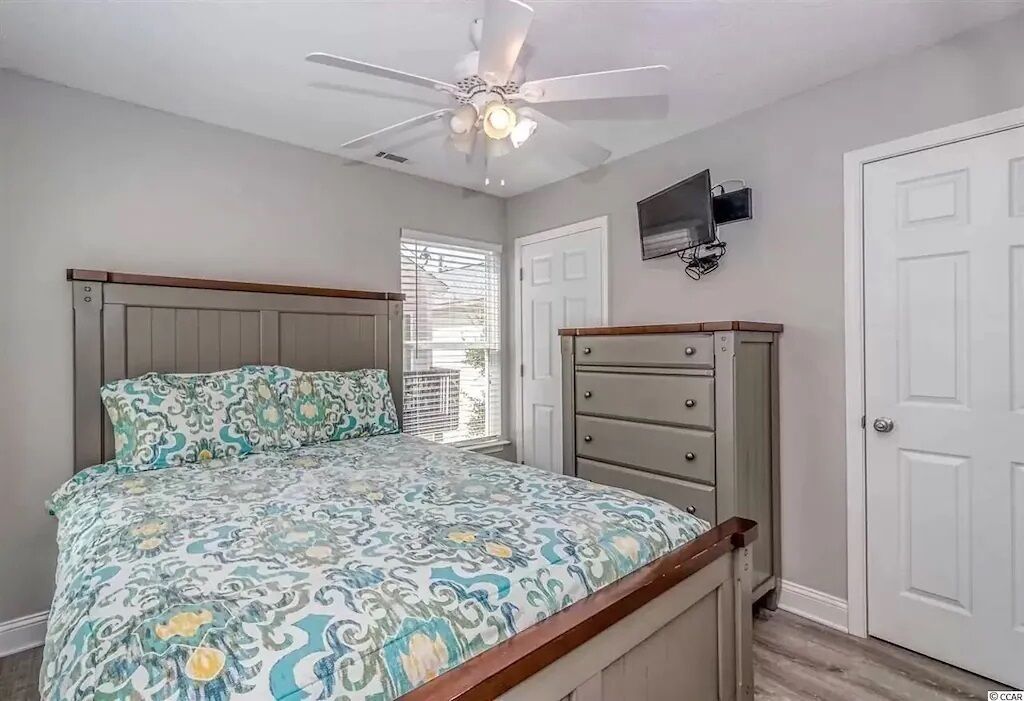 A bedroom with a bed, a dresser, a ceiling fan, and a TV mounted on the wall.