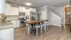 A kitchen with white cabinets, a wooden island with stools, stainless steel appliances, and a staircase leading to another level. - Welcome to Serinity on the Beach (Myrtle Beach)
