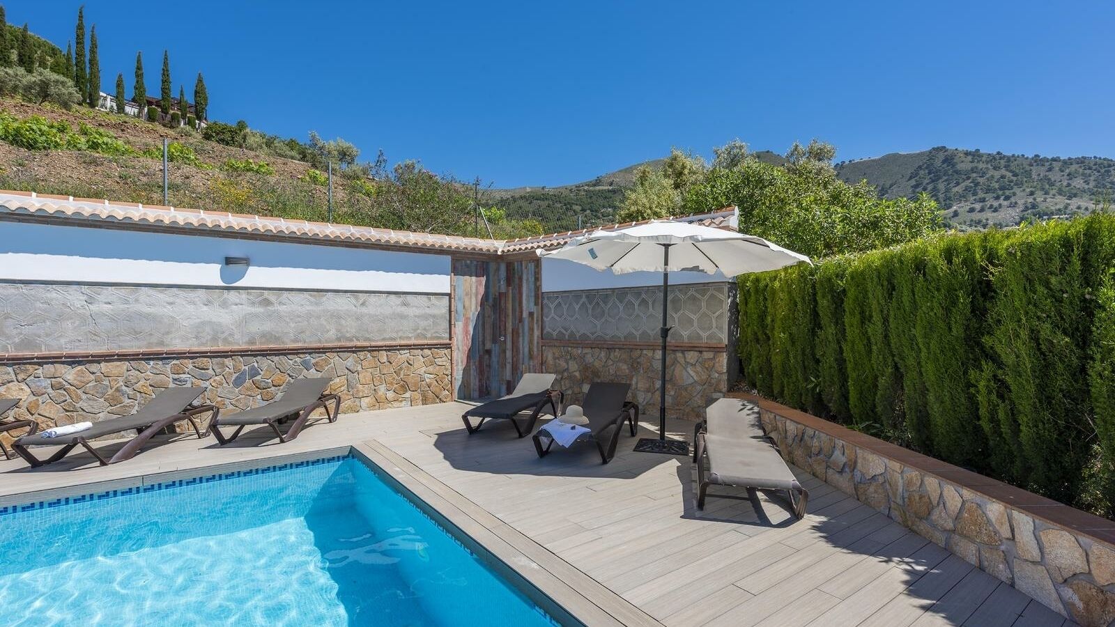Ein Pool mit Liegestühlen und Sonnenschirm, umgeben von Steinmauern und Blick auf Hügel.