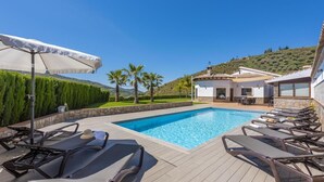Un espace aménagé au bord de la piscine, avec des chaises longues et un parasol, un mur en pierre et un bâtiment doté d’un toit en tuiles.