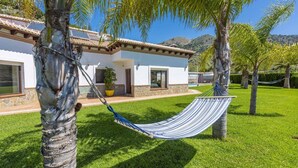 Une maison à l’extérieur blanc, avec des fondations en pierre et un toit recouvert de tuiles. Un hamac est suspendu entre deux arbres dans le jardin.