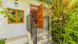 Un escalier menant à une porte en bois ornée d’une rampe métallique, le tout entouré d’une végétation luxuriante.