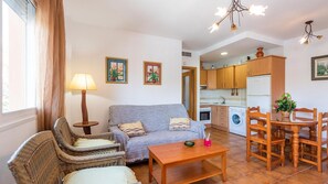 A living room with a sofa, two armchairs, a wooden coffee table, and a dining table with chairs. There is a kitchen area with wooden cabinets and a washing machine. - Apartamento Cañada del Barco Torrox by Ruralidays (Málaga)