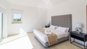 Ein modernes Schlafzimmer mit einem großen Bett, einem Nachttisch mit Lampe und Blick ins Freie durch ein Fenster.