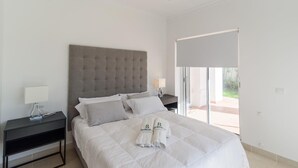 A modern bedroom with a large bed, a grey tufted headboard, two bedside tables with lamps, and a sliding glass door leading to an outdoor area. - Villa Latina Golf Arcos de la Frontera by Ruralidays (Cádiz)