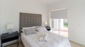 A modern bedroom with a large bed, a grey tufted headboard, two bedside tables with lamps, and a sliding glass door leading to an outdoor area. - Villa Latina Golf Arcos de la Frontera by Ruralidays (Cádiz)